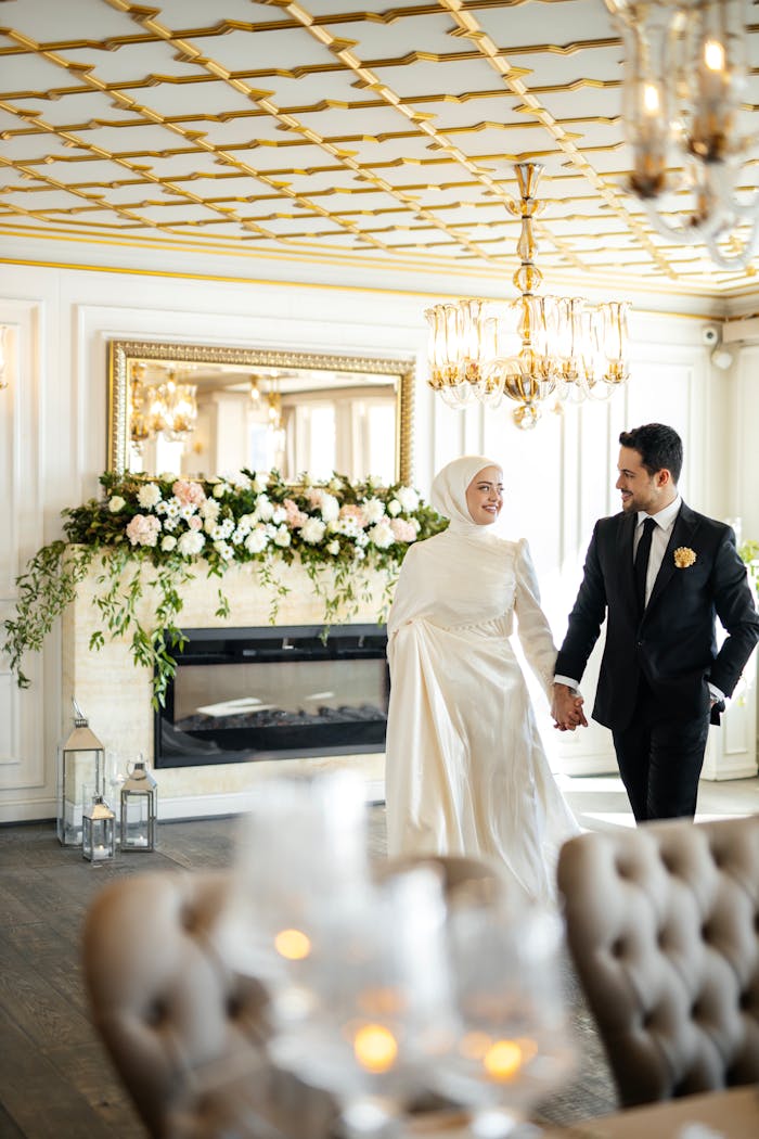 A Muslim bride and groom in a luxurious room with floral decor, smiling.