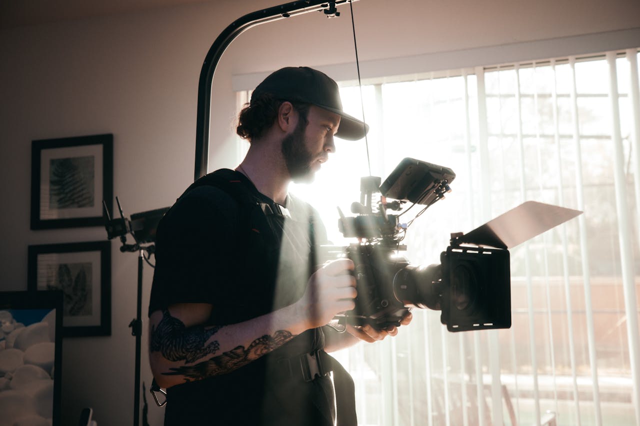 A bearded videographer in a black shirt operates a film camera inside a sunlit room, showcasing filmmaking in action.
