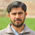 Close-up portrait of a bearded man in a field, looking directly at the camera.